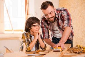 precisión y perfección. joven carpintero sonriente mostrando a su hijo cómo lijar madera en su taller foto