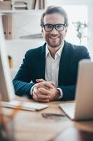 Ready to hear you out. Handsome young man in eyeglasses smiling and looking at camera while sitting on working place in creative office photo