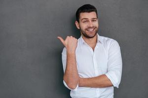 Pointing copy space. Handsome young man pointing copy space and smiling while standing against grey background photo