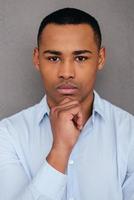 Portrait of confidence. Confident young African man holding hand on chin and looking at camera while standing against grey background photo