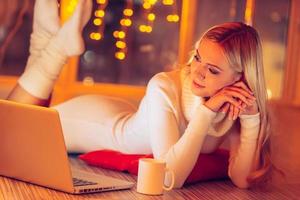 Home is the best place in the world. Beautiful young woman in white sweater and socks lying on the floor and looking at laptop with Christmas lights in the background photo