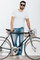 Just me and my bike. Full length of handsome young Indian man in sunglasses leaning at his retro styled bicycle while standing against white background photo