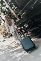 Confident businesswoman. Full length rear view of young woman in suit pulling luggage while walking outdoors photo