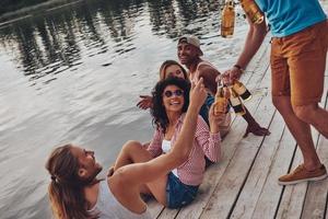 hora de refrescarse. grupo de jóvenes felices con ropa informal sonriendo y bebiendo cerveza mientras se sientan en el muelle foto