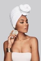 Dangerously beautiful. Attractive young African woman in turban adjusting earring and looking away while standing against grey background photo