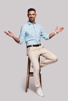 Greeting. Full length of good looking young man smiling and keeping arms outstretched while sitting on stool against grey background photo