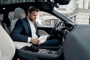 Checking the schedule. Handsome young man in full suit holding his personal organizer and looking at it while sitting inside of the car photo