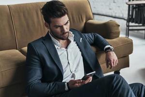 Important message. Handsome young man in full suit holding a smart phone and looking away while sitting on the floor at home photo