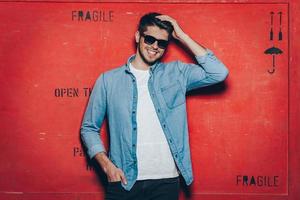 Cheerful and trendy.Handsome young cheerful man in sunglasses keeping hand in hair and looking at camera with smile while standing against red background photo