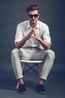 Mr Cool. Full length of confident young handsome man in sunglasses keeping hands clasped and looking at camera while sitting in chair against grey background photo