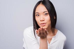 Planting a kiss on your heart. Beautiful young Asian woman sending a kiss at camera while standing against grey background photo