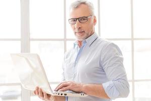 Supporting your business. Confident grey hair senior man in shirt working on laptop and looking at camera while standing in front of the window photo