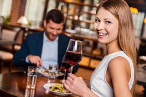 Enjoying time together. Beautiful young woman holding glass with red wine and smiling while sitting at the restaurant with her boyfriend sitting in the background photo