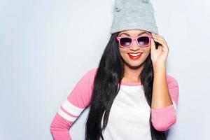 Stylish beauty. Happy young African woman in funky clothes adjusting her sunglasses and smiling while standing against grey background photo