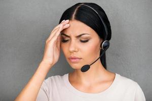 Stressful day at work. Frustrated young female customer service touching her head and keeping eyes closed while standing against grey background photo