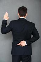 Telling lies. Rear view of young businessman keeping his fingers crossed and arm raised while standing against grey background photo