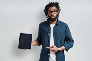 Playful young African man in eyewear pointing copy space using digital tablet while standing against grey background photo