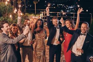 grupo de personas hermosas en ropa formal sosteniendo bengalas y sonriendo mientras pasan tiempo en una fiesta al aire libre foto