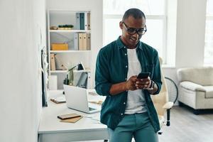 Using new mobile app. Handsome young African man in shirt using smart phone while standing in the office photo