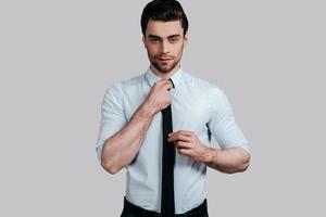 Used to look perfect.  Good looking young man in formalwear looking at camera and adjusting his tie while standing against grey background photo