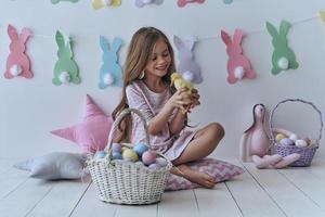 ¿Estás listo para celebrar a una niña linda jugando con un pato de juguete y sonriendo mientras te sientas en la almohada con decoración en el fondo? foto