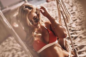 simplemente relajarse. atractiva mujer joven en bikini rojo ajustando la flor en su cabello mientras se sienta en una hamaca en la playa foto