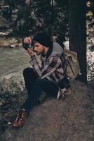 capturando la belleza. joven moderno con mochila fotografiando la vista mientras se sienta en el bosque con el río de fondo foto