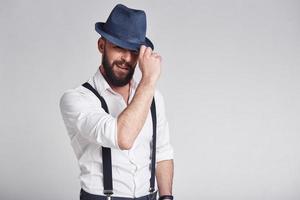 Charming gangster. Handsome young man in suspenders adjusting his hat and looking at camera with smile while standing against grey background photo