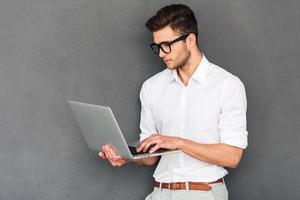 Checking his timetable. Confident young man holding laptop and typing while standing against grey background photo