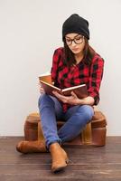 Creating a comfort around her.  Beautiful young woman in headwear holding a book and cup of coffee while sitting on suitcase photo