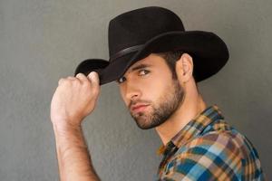 Wild West is his heart. Handsome young man adjusting his cowboy hat and looking at camera while standing against grey background photo