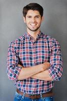 Confident handsome. Smiling young man keeping arms crossed and looking at camera while standing against grey background photo