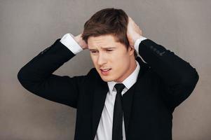 Failed again. Frustrated young man in formalwear touching head with hands while standing against grey background photo