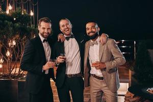 Three cheerful men in suits holding whiskey glasses and looking at camera while spending time on party photo