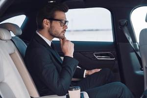 Thinking about solution. Thoughtful young man in full suit keeping hand on chin while sitting in the car photo