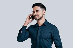 Pleasant business talk. Good looking young man talking on smart phone and looking away while standing against grey background photo