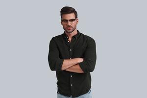 Serious look. Thoughtful young man in eyeglasses keeping arms crossed and looking at camera while standing against grey background photo