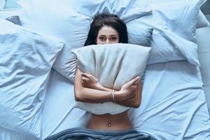 I want to stay in bed. Top view of attractive young woman covering half of her face with pillow and looking at camera while lying on the bed at home photo