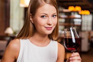 belleza con vino. bella joven sosteniendo un vaso con vino tinto y sonriendo mientras se sienta en el restaurante foto