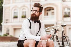 Enjoying city life. Low angle view of young bearded man in eyewear holding mobile phone and looking away while sitting outdoors photo
