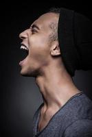 Energy inside him. Side view of furious young African man in beanie keeping eyes closed and mouth open while standing against black background photo