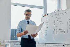 Confident English teacher going through papers while standing near the whiteboard at classroom photo