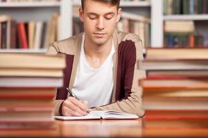 Doing his research in library. Confident young man writing something in his note pad while sitting at the library desk photo
