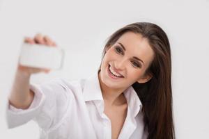 belleza con teléfono móvil. bella joven fotografiando con la cámara de su teléfono móvil y sonriendo mientras está aislada en blanco foto