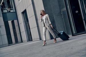 Organizing every minute. Full length of young woman in suit pulling luggage and smiling while walking outdoors photo