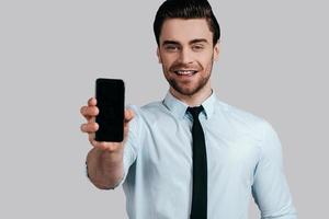 Copy space on his mobile phone. Good looking young man in white shirt and tie showing a smart phone and looking at camera while standing against grey background photo