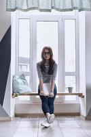 Young and beautiful. Attractive young woman holding a notebook and looking at camera while sitting on the window sill photo