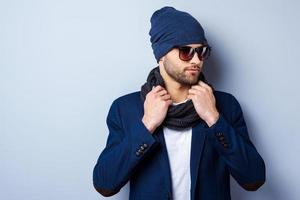 en su propio estilo. apuesto joven elegante con gafas de sol y sombrero ajustando su bufanda y mirando hacia otro lado mientras está de pie contra un fondo gris foto