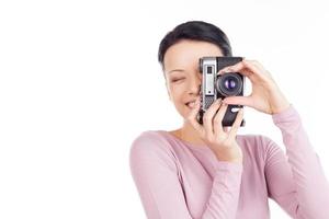 She likes photographing. Beautiful young woman focusing at you with camera and smiling while isolated on white photo