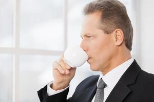 Break time. Side view of mature man in formalwear drinking coffee and looking through a window photo